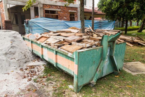 A large, rectangular green metal skip filled with a substantial pile of wooden debris, including planks and panels of varying sizes, some with uneven edges and rough finishes. The skip is positioned on a patch of earth and grass, with a concrete pathway nearby. It is situated outdoors in a residential or semi-urban environment, with a mound of light-colored construction waste or rubble visible to the left side of the skip. In the background, there is a partially constructed brick building covered with a blue tarpaulin, supported by wooden poles, and some mature trees casting shadows on the ground. The scene is outdoors during daylight with natural light illuminating the area, reflecting a typical rubbish removal scenario managed by Commercial Waste Removal Camden Town, highlighting the handling of building or renovation waste in the local area.