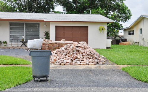 In front of a residential property on a driveway, there is a large pile of light pinkish-beige bricks stacked haphazardly, with some scattered on the dark asphalt surface. To the left of the brick pile, a grey wheelie bin is positioned on the edge of the driveway, partially covering the lower part of the brick stack. The background shows the front of a single-story house with a light-colored exterior, a large window with horizontal blinds, and a brown garage door. On the right side, part of another similar house is visible, along with some outdoor clutter. Green grass borders the driveway on both sides, with a small tree or shrub hanging over the right side of the garage. The overall scene suggests a rubbish removal or site clearance activity, involving the collection and disposal of construction debris, likely managed by companies such as Commercial Waste Removal Camden Town.
