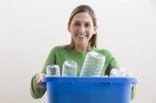 A young woman with a cheerful expression, wearing a green sweater, holding a blue recycling bin filled with clear plastic bottles of various sizes and shapes, some with ridges and caps, in an indoor setting with a plain white or light background. The bottles appear clean and are arranged upright within the bin. The image illustrates the collection or disposal of recyclable plastic waste, relevant to rubbish removal services offered by Commercial Waste Removal Camden Town, which operates in the Camden Town area including postcodes around NW1. The environment is simple and uncluttered, focusing on the waste items and the bin, with natural lighting highlighting the transparency and textures of the plastic bottles. The scene emphasizes responsible waste management and the importance of recycling in line with professional rubbish removal practices.