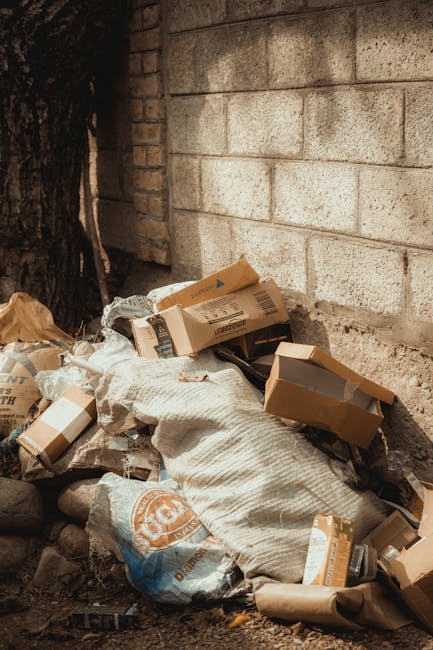 A close-up view of a large, weathered, beige cloth bag filled with discarded plastic bottles and other waste materials, positioned on a narrow urban street pavement next to a graffiti-marked wall with